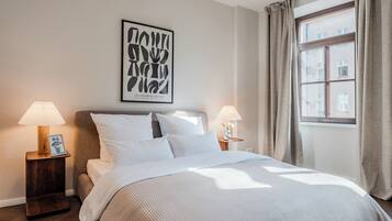 A bedroom with a bed, two bedside tables with lamps, a framed artwork on the wall, and a window with curtains.