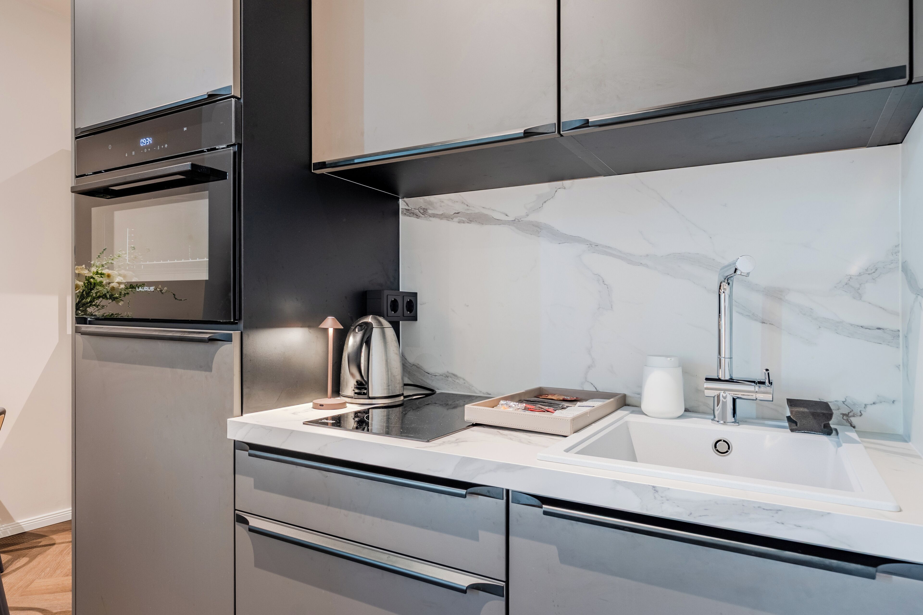 Una cocina moderna con horno integrado, hervidor y lavamanos con salpicadero de mármol.