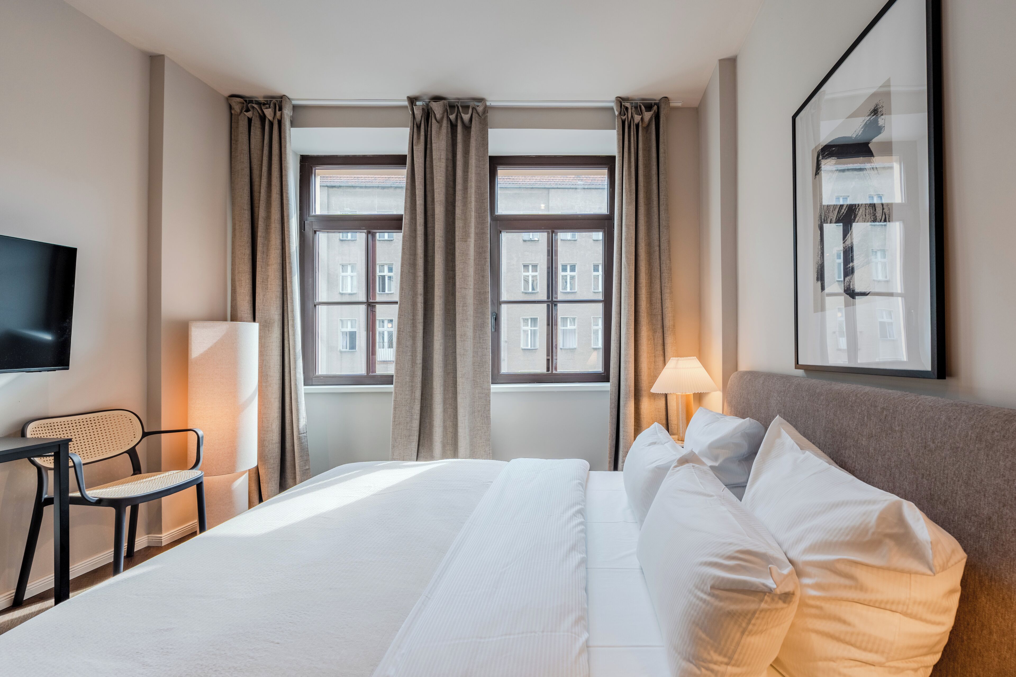 Et hotelværelse med seng, stol, skrivebord, lampe, fjernsyn og et vindue med gardiner.