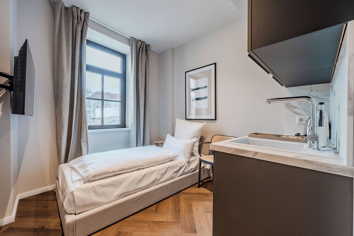 Habitación de hotel moderna con cama, ventana con cortinas, televisor montado, cuadro enmarcado en la pared y baño con lavamanos y espejo.