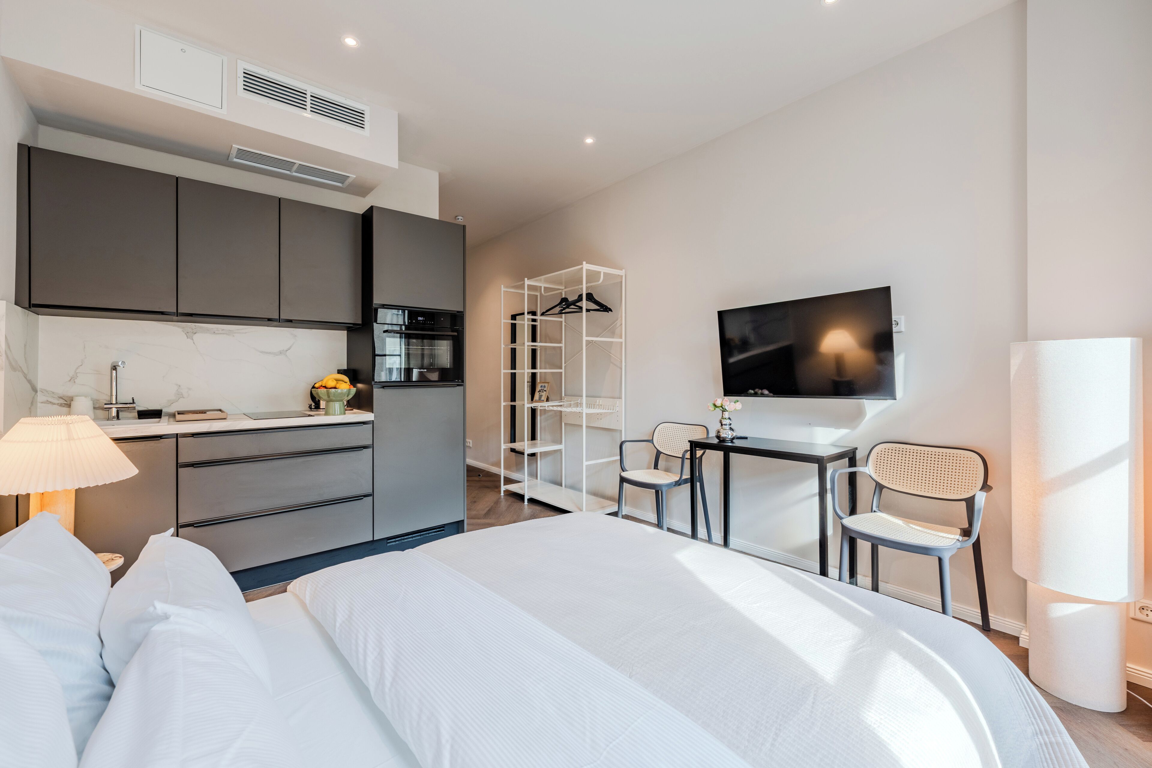 A modern hotel room with a bed, a desk with a chair, a TV, a kitchenette, and a wardrobe.