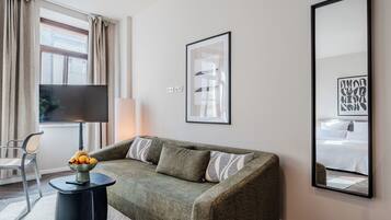 Una habitación de hotel moderna con un sofá verde, una mesita de noche con frutero, un televisor de pantalla plana y un espejo grande que refleja una cama y una obra de arte enmarcada.