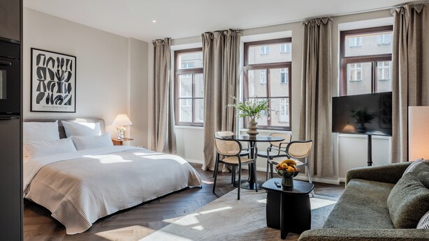 Et hotelværelse med seng, spisebord med stole, sofa, fjernsyn og store vinduer med gardiner.