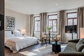 Une chambre d’hôtel comprenant un lit, une table à manger avec des chaises, un canapé, un téléviseur et de grandes fenêtres avec des rideaux.