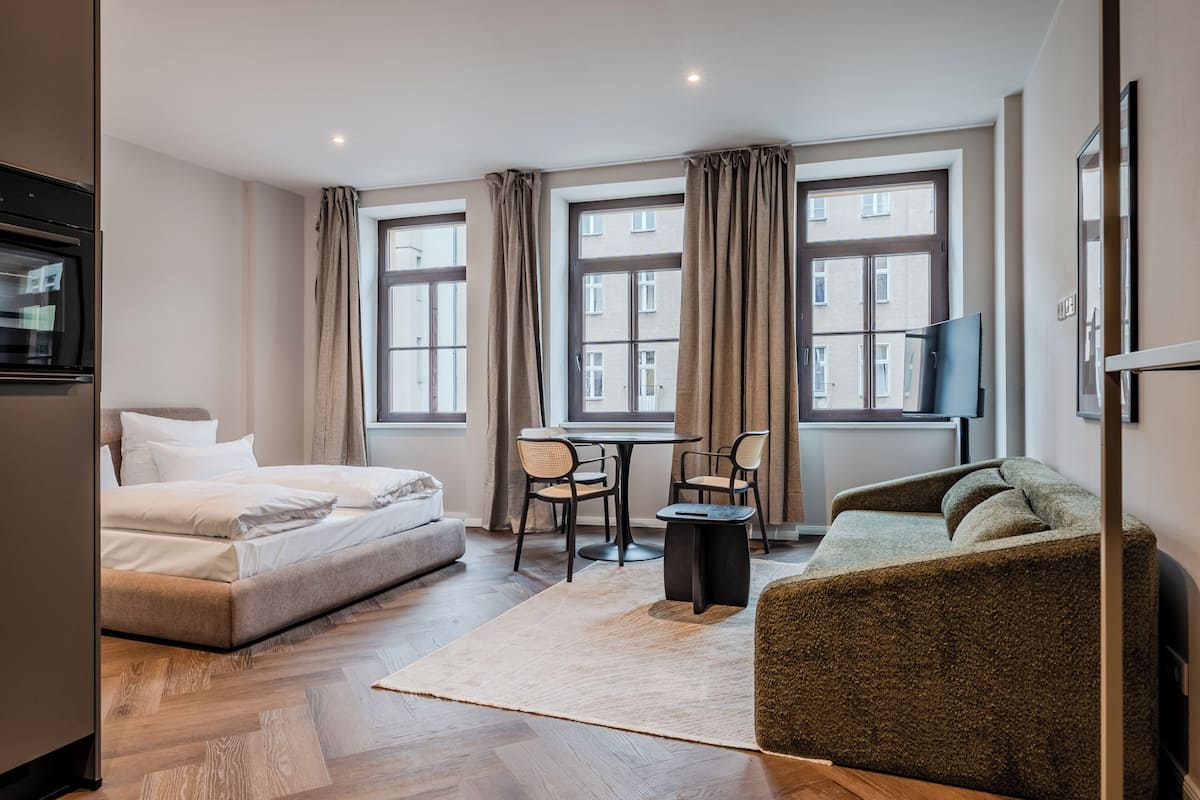 Habitación de hotel con cama, sofá, mesa de comedor con sillas, televisión y ventanas grandes con cortinas.