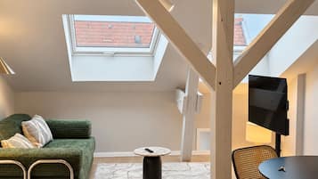 Une chambre de style loft moderne, dotée d’une lucarne, d’un canapé vert, d’une table basse ronde et d’un téléviseur fixé au mur.