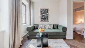En stue med en grøn sofa, et sofabord med frugt, et indrammet abstrakt kunstværk og udsigt til et andet rum.