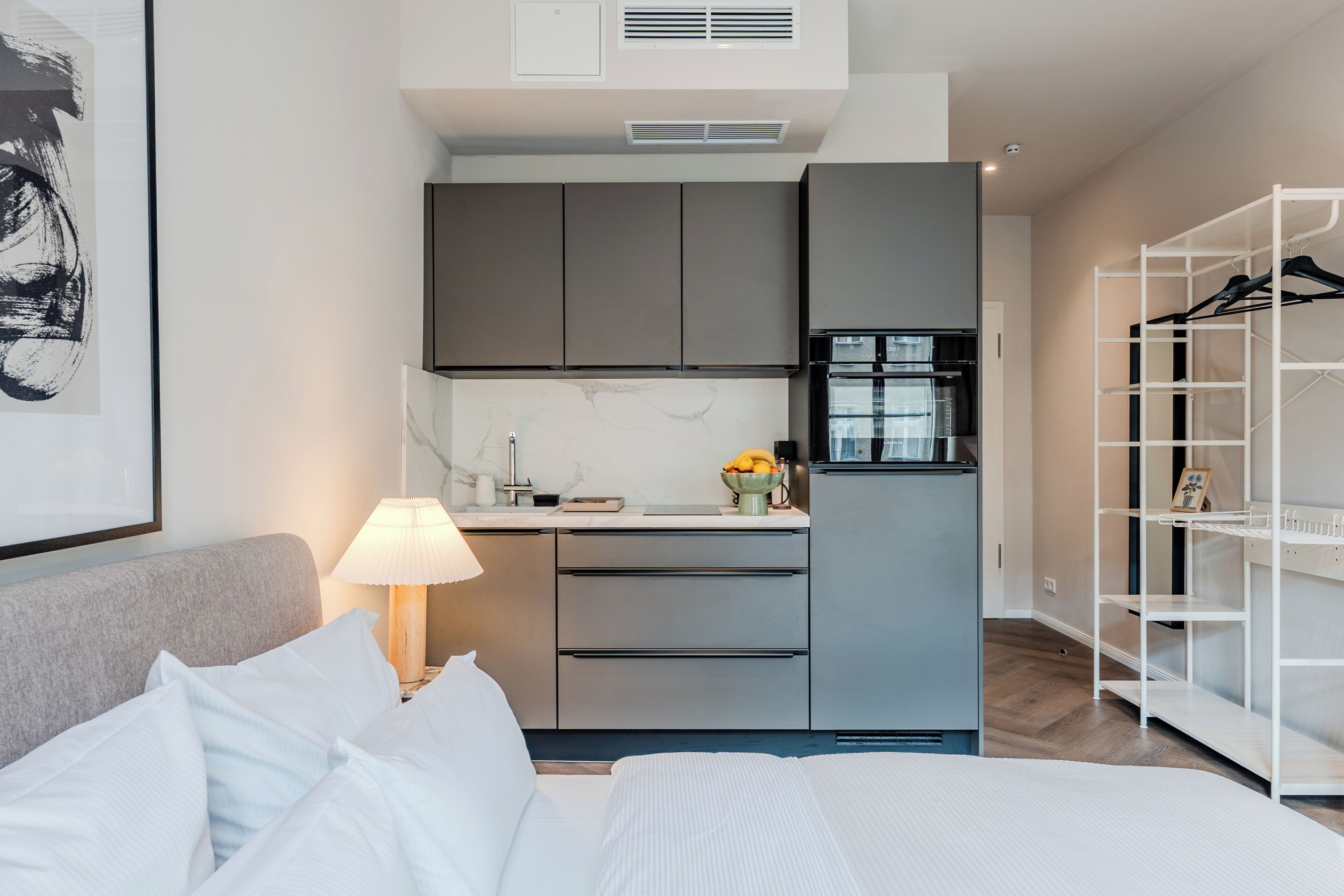 Moderni hotellihuone, jossa on sänky, yöpöytä lampun kanssa, pieni keittiö jääkaapilla ja vaatekaappi.