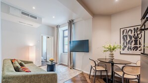 Ein modernes Wohnzimmer mit Sofa, einem Esstisch mit Stühlen, einem flachbildfernseher an der Wand und einem Fenster mit Vorhängen.