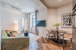 Ein modernes Wohnzimmer mit Sofa, einem Esstisch mit Stühlen, einem flachbildfernseher an der Wand und einem Fenster mit Vorhängen.