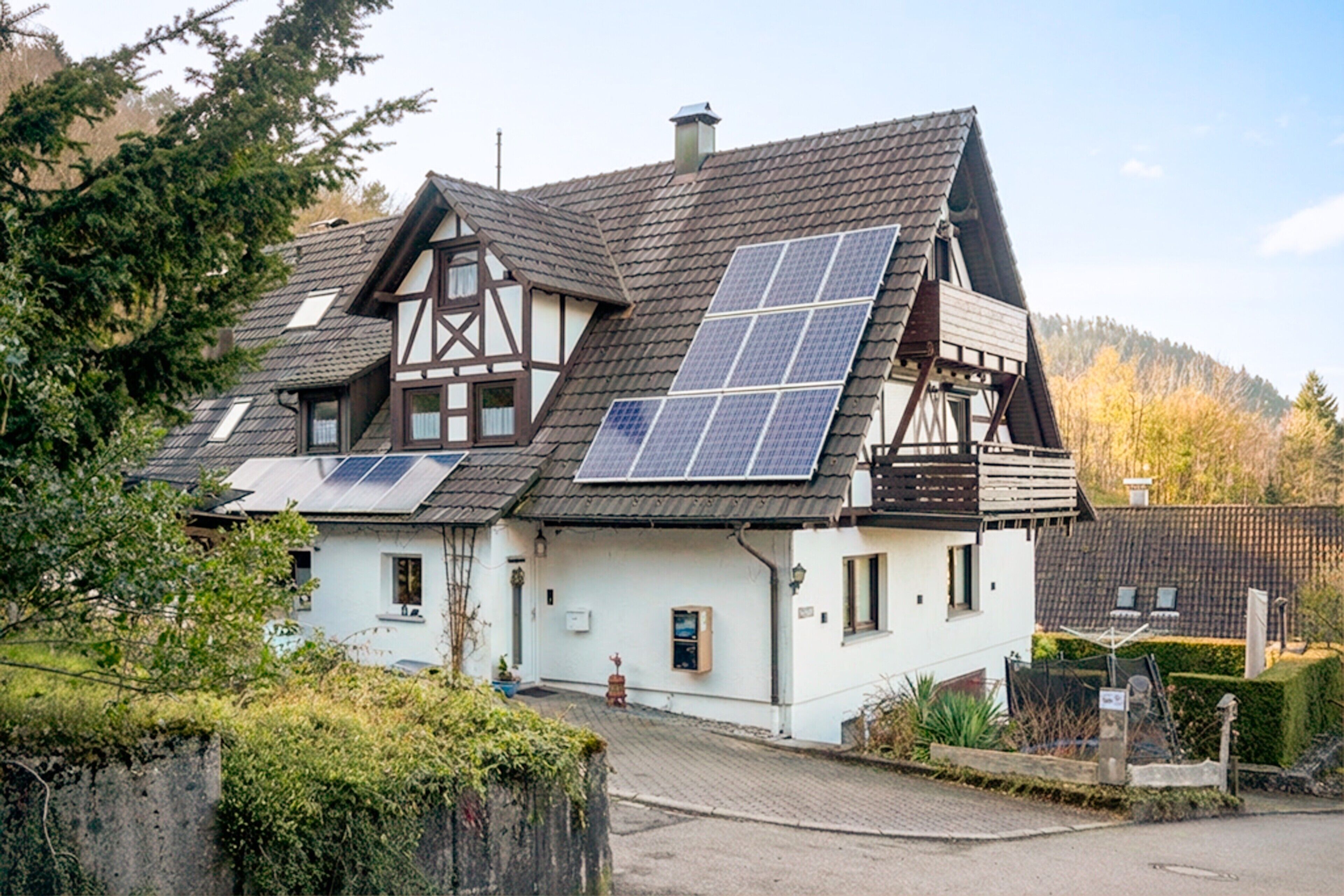 Ein traditionelles Fachwerkhäuschen mit Solaranlage auf dem Dach, umgeben von viel Grün und einer gepflasterten Auffahrt.