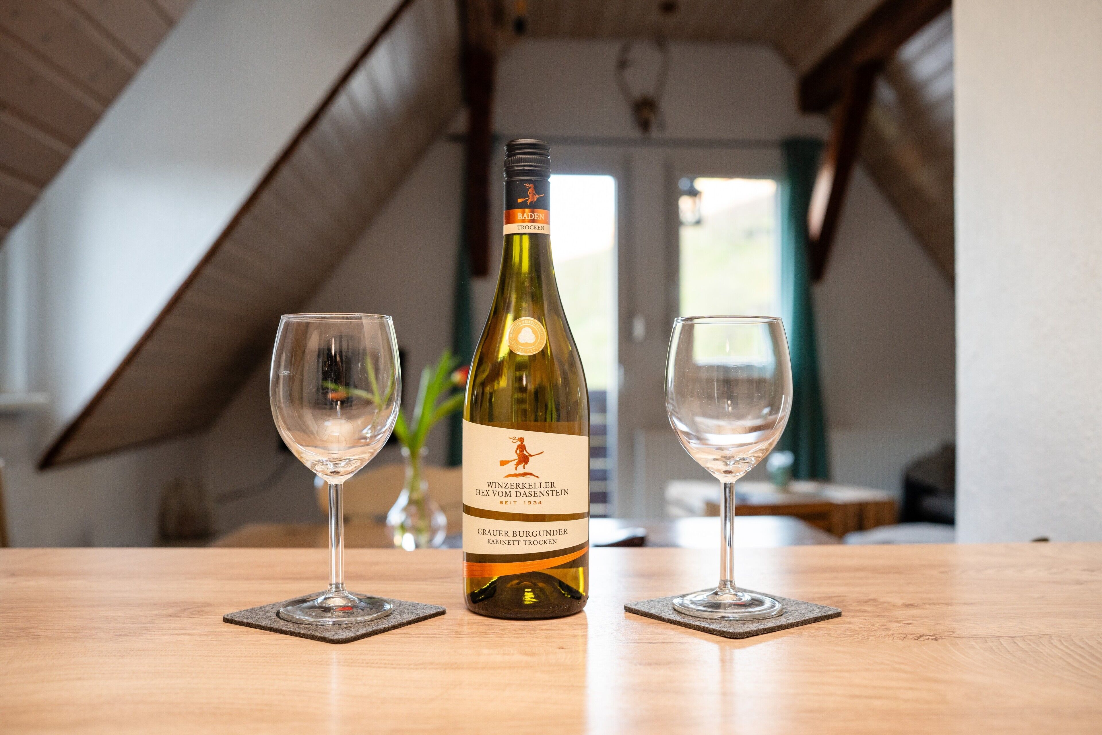 Een fles wijn en twee glaasjes op een houten tafel in een kamer met schuine plafonds.