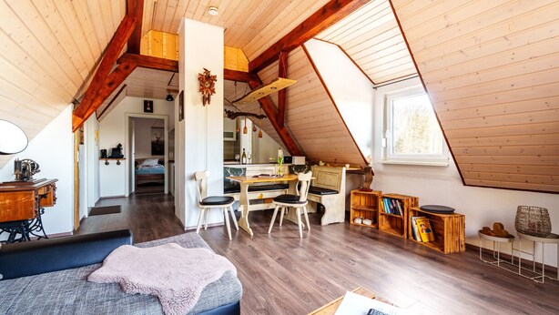 Ein Loft-Zimmer mit schrägverdachter Decke, Holzbalken, einem Essbereich mit Tisch und Stühlen, einer Nähmaschine und einer gemütlichen Sitzecke mit Sofa und Decke.