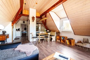 Ein Loft-Zimmer mit schrägverdachter Decke, Holzbalken, einem Essbereich mit Tisch und Stühlen, einer Nähmaschine und einer gemütlichen Sitzecke mit Sofa und Decke.