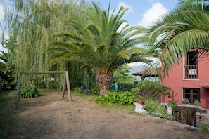 A garden with a swing set, palm trees, and a red building with a window. - Rural House 'Casa Remis' in Piloña – Sleeps 12, Private Bathrooms, Fireplace (Piloña)