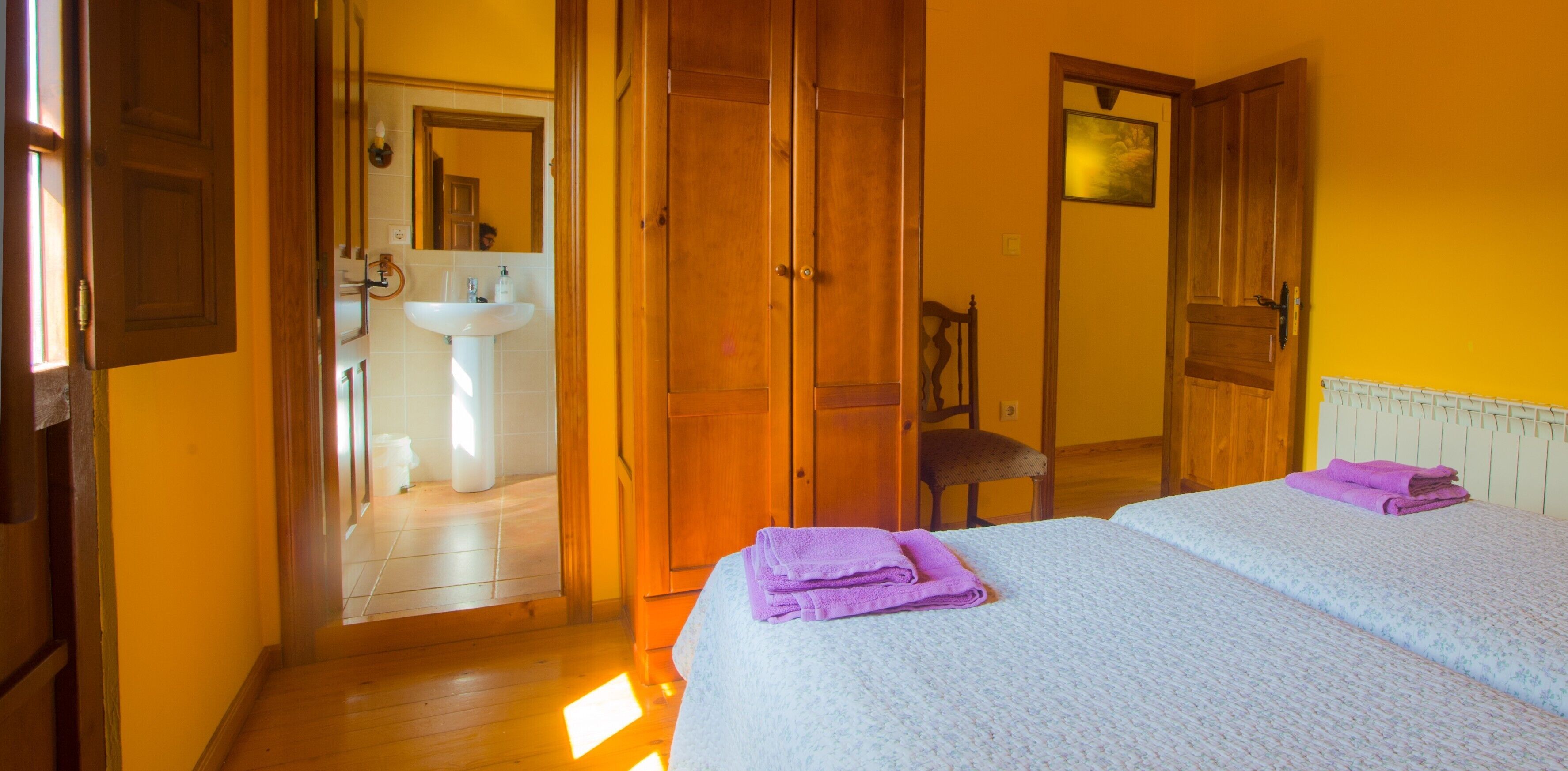 Een kamer met een bed, een stoel, een houten kledingkast en een badkamer met een wastafel en spiegel.