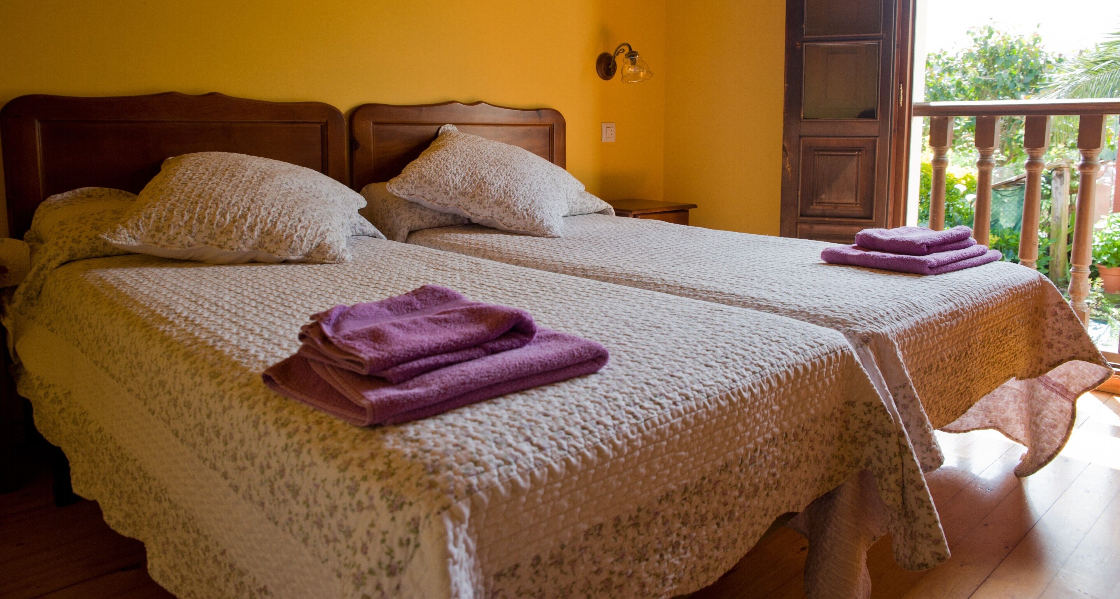 A double bed with a floral-patterned bedspread and two pillows, a wooden nightstand, and a balcony with a wooden railing.