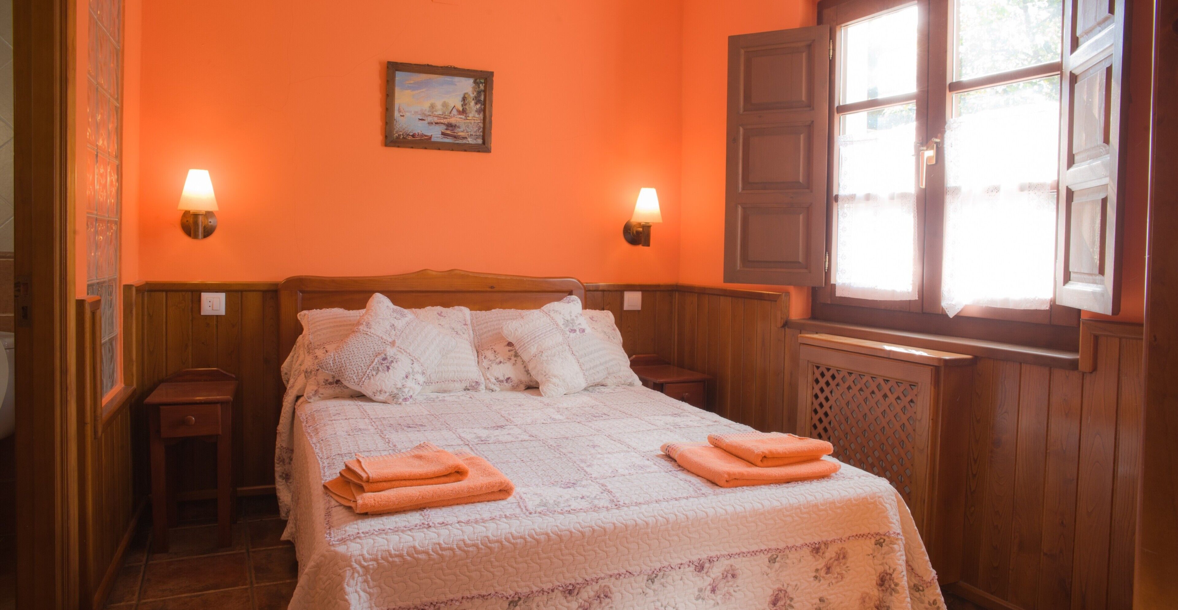 Une chambre à coucher avec une tête de lit en bois, deux tables de chevet, un lit recouvert d’une courtepointe, deux lampes fixées au mur et une fenêtre avec des volets.