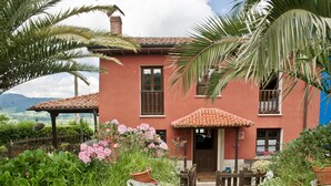 A red house with a tiled roof, surrounded by lush greenery and flowering plants. - Rural House 'Casa Remis' in Piloña – Sleeps 12, Private Bathrooms, Fireplace (Piloña)