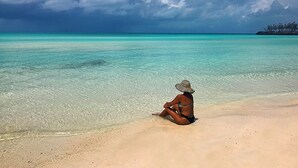 A person sitting on a sandy beach with clear turquoise water and a cloudy sky. - Brand New Nov 2025, Affordable. Great Location, Right Next to Airport Beach. (Governors Harbor)