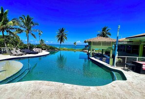 A pool with a curved edge, lounge chairs, and a thatched-roof structure. - Brand New Nov 2025, Affordable. Great Location, Right Next to Airport Beach. (Governors Harbor)