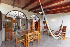 A spacious dining area with a wooden table and chairs, a hammock, and a view of the kitchen and living area. - Villas el Parque 18A: Ocean-View Studio Apartment, Central, Pool, AC & WiFi (Quepos)