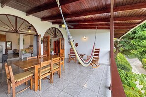A spacious dining area with a wooden table and chairs, a hammock, and a view of a lush garden. - Villas el Parque 18A: Ocean-View Studio Apartment, Central, Pool, AC & WiFi (Quepos)