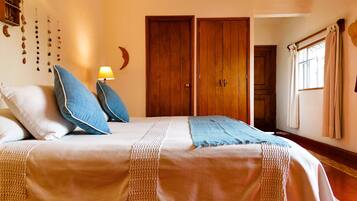 Un dormitorio con cama, almohadas, mesita de noche, lámpara, armario y ventana con cortinas.