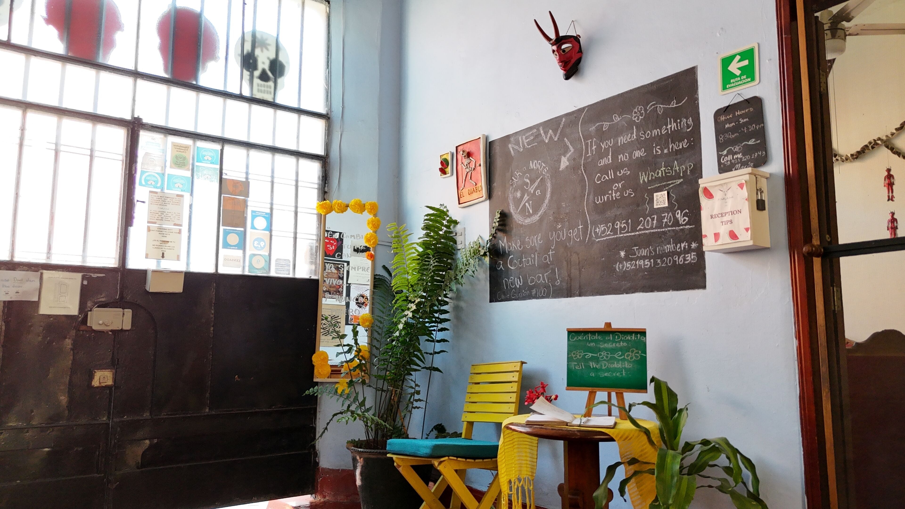 Una habitación con ventana, puerta, una pizarra con escritura, una mesita con una planta y una silla.