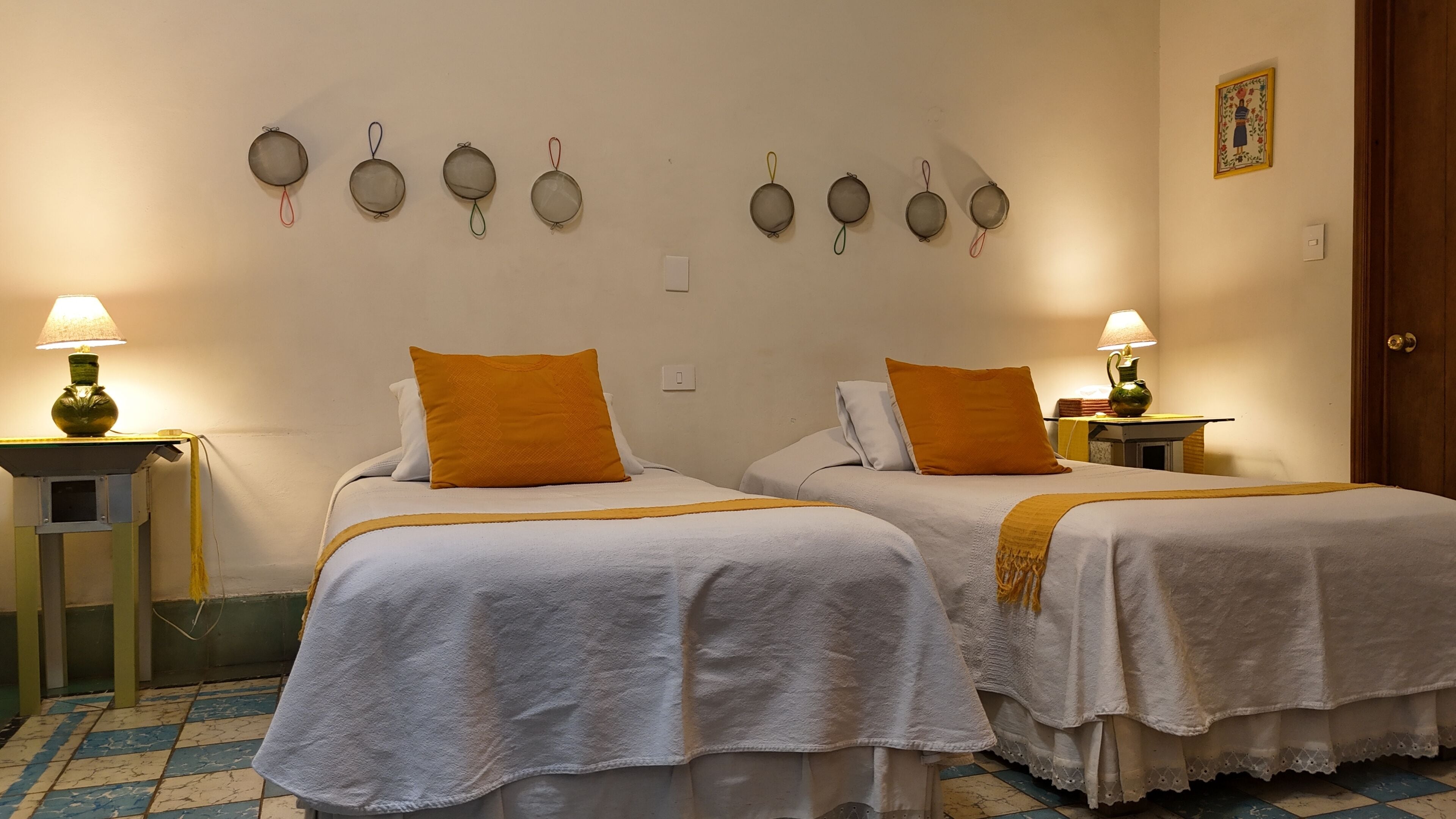 A hotel room with two beds, each with a white bedspread and an orange pillow. There are wall-mounted lamps and a small table with a lamp.