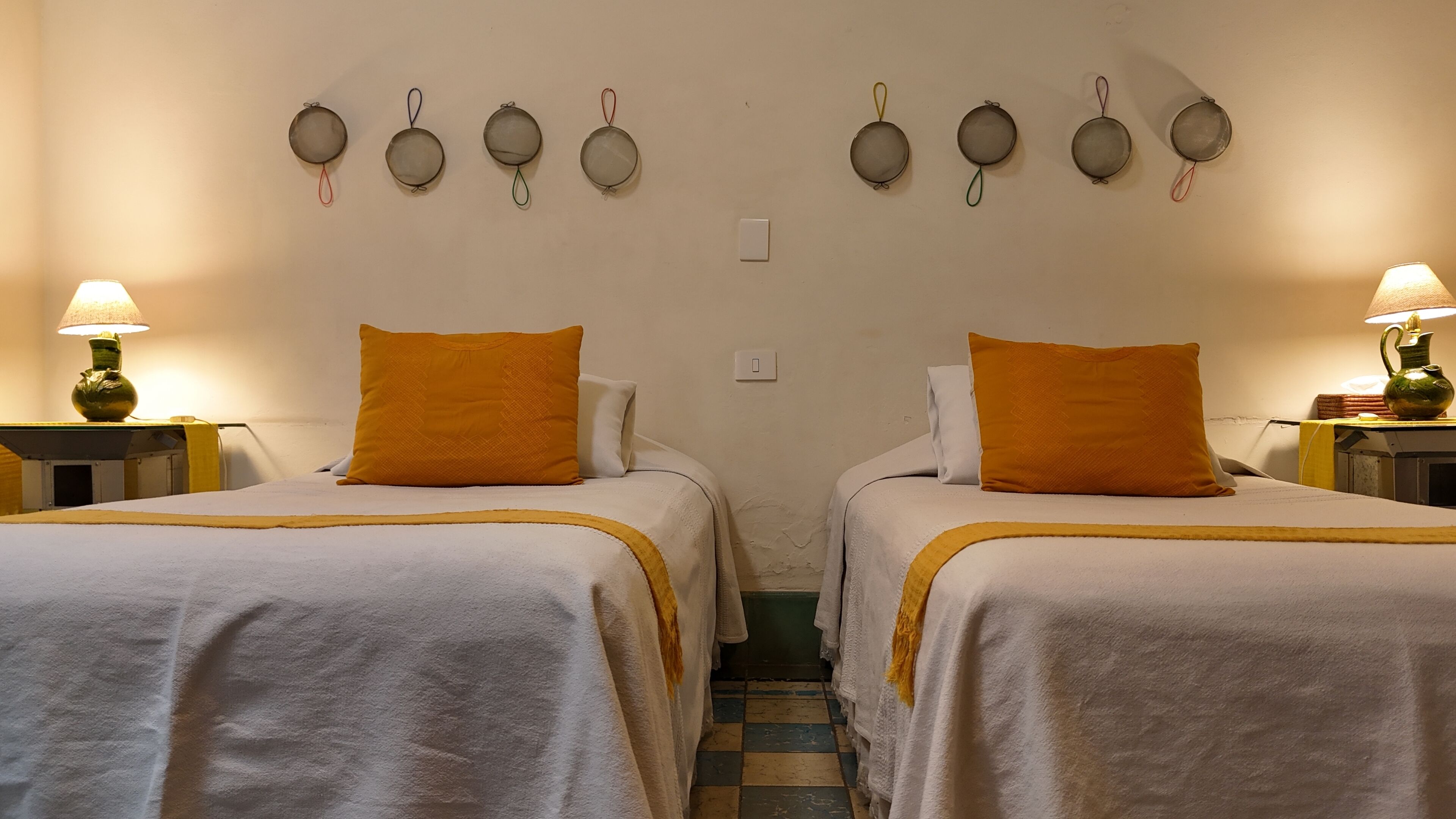 A hotel room with two beds, each with a white bedspread and an orange pillow. There are wall-mounted light fixtures above the beds and a small white switch on the wall.