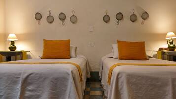 Habitación de hotel con dos camas, cada una con cubrecama blanco y almohada naranja. Hay lámparas fijadas a la pared sobre las camas y un interruptor blanco pequeño en la pared.