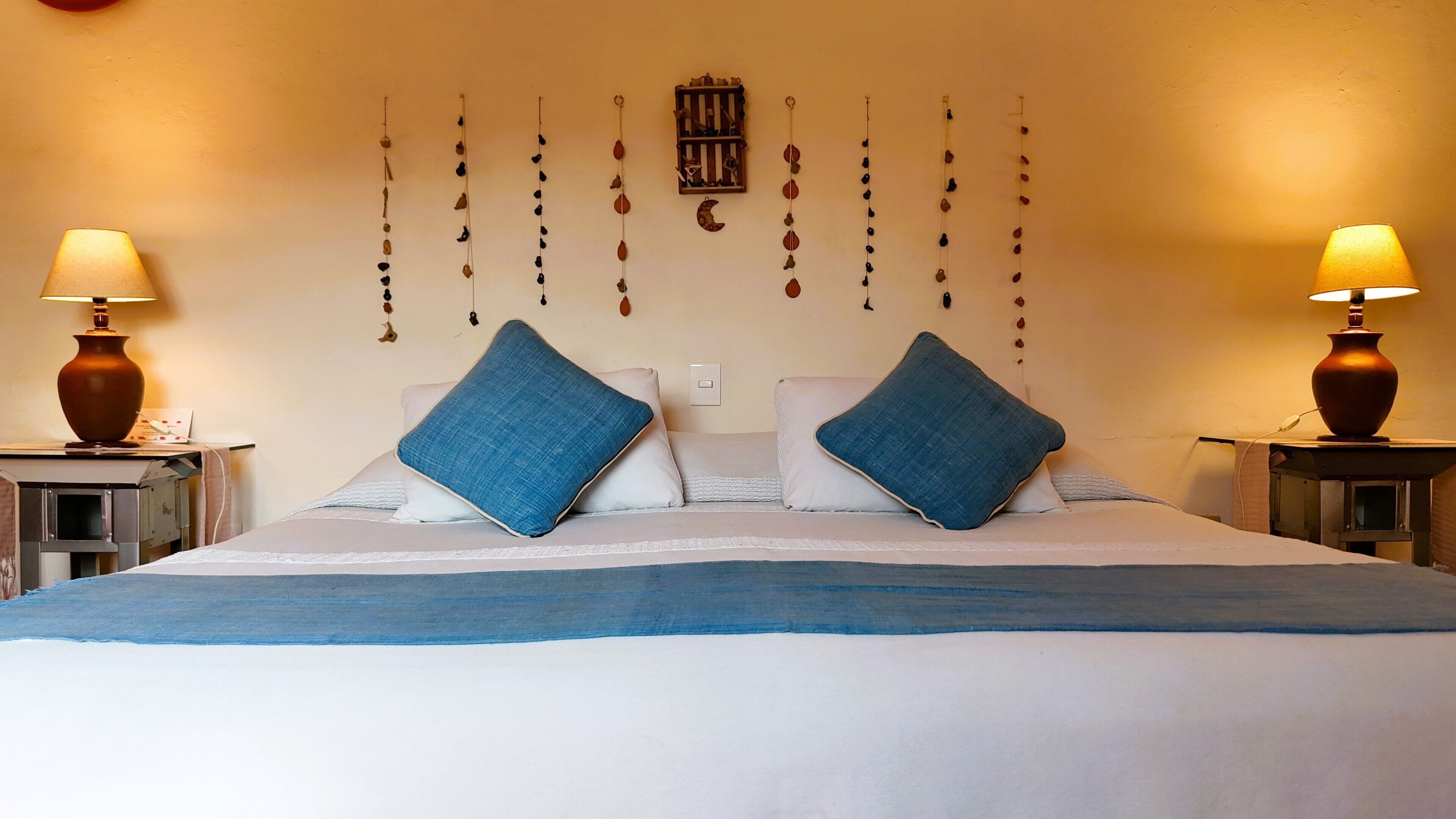 A neatly made bed with blue pillows, two bedside lamps, and a wall-mounted light fixture with hanging decorations.