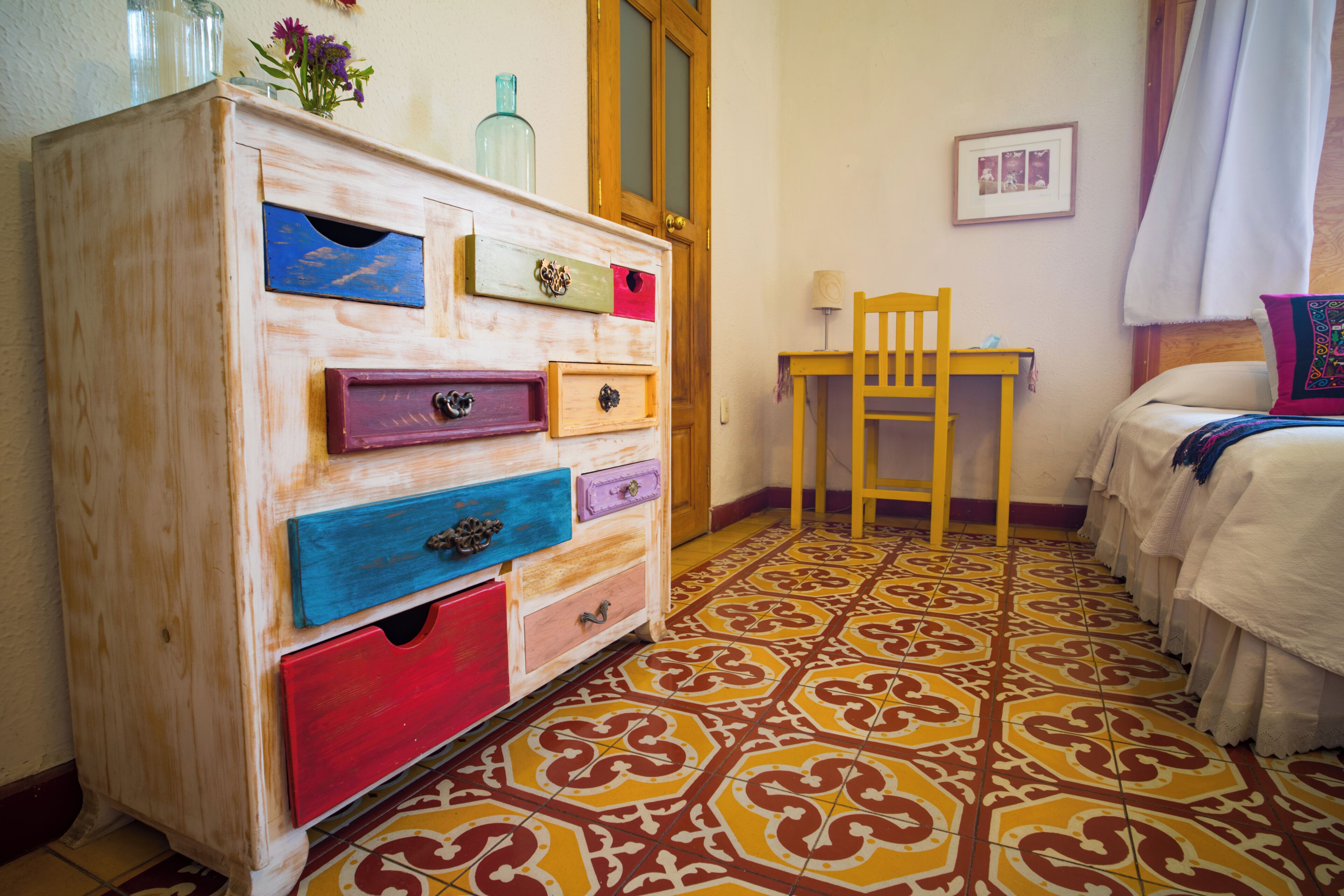 A room with a wooden dresser with multiple drawers, a chair, a bed with white bedding, and a patterned floor.