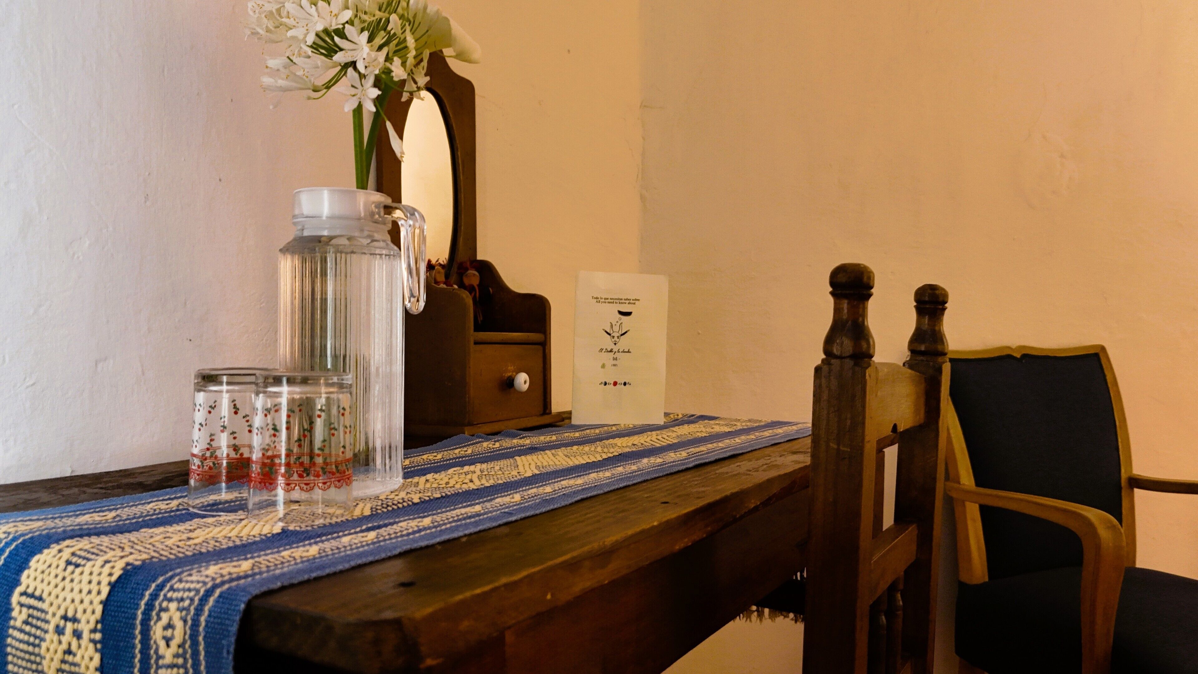 A wooden table with a blue and white patterned tablecloth, a clear glass pitcher, and a small vase with white flowers.