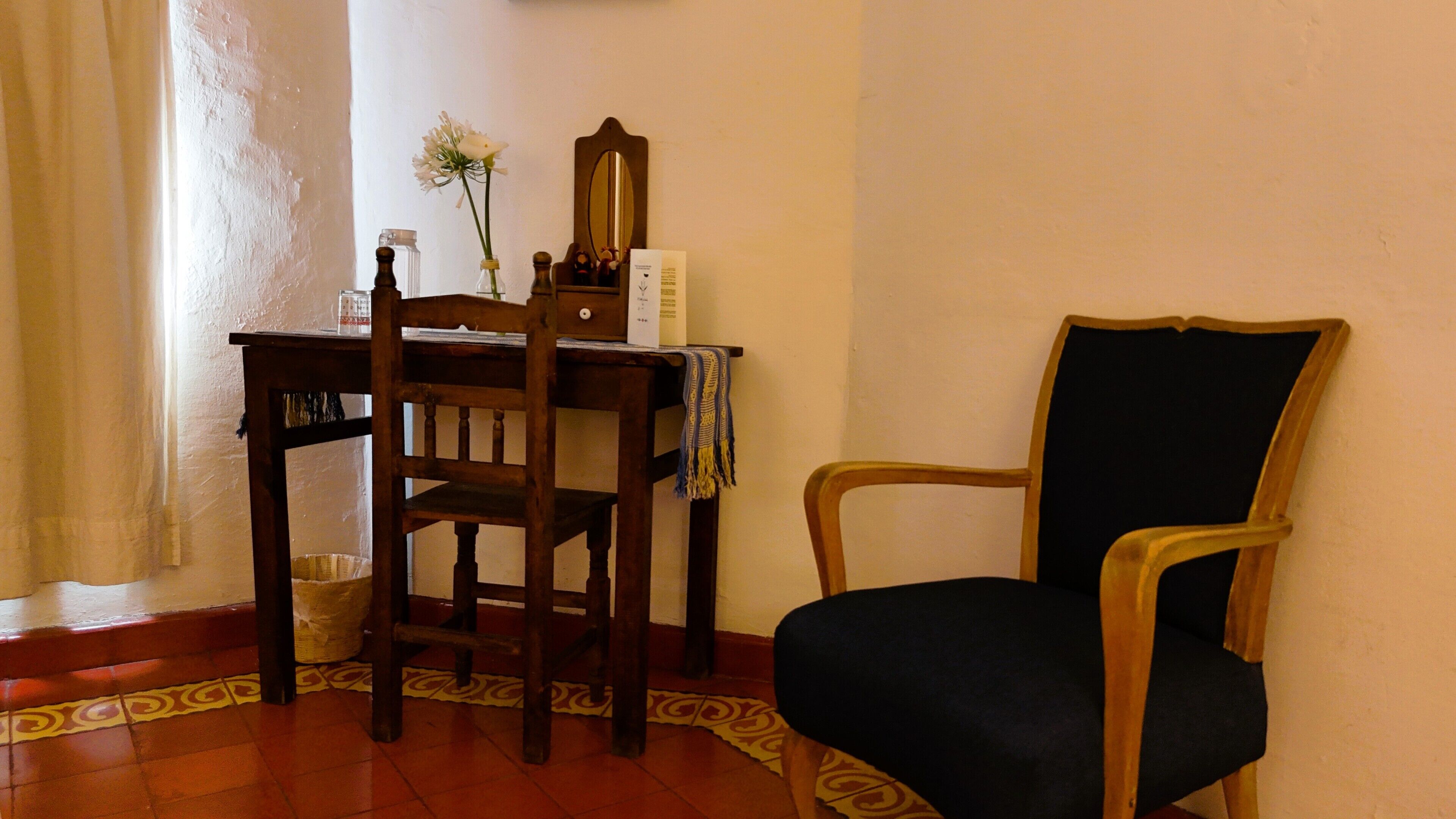 A wooden desk with a chair, a vase with a flower, a mirror, and a small table with a box and a candle.