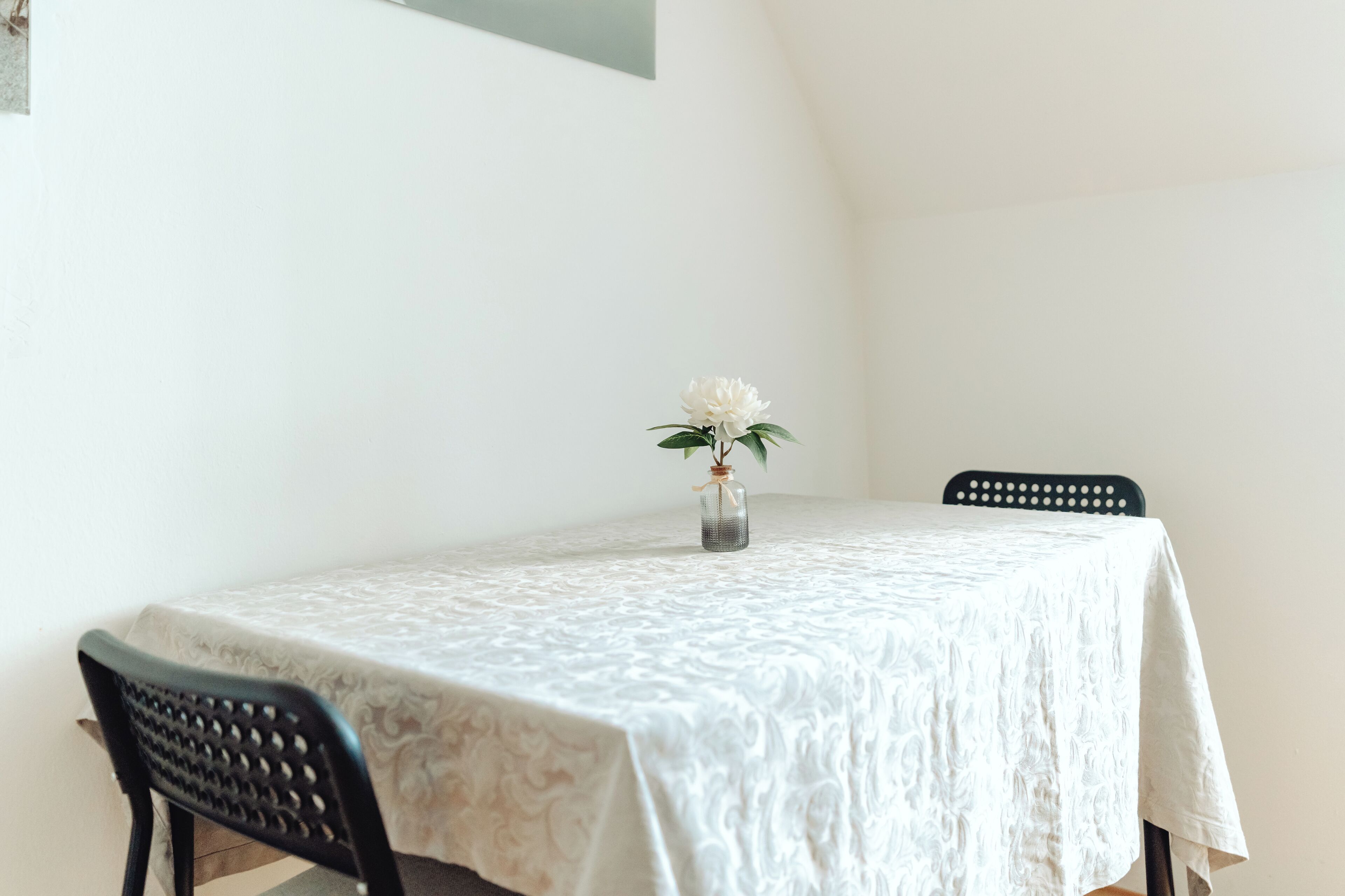 A dining table with a white tablecloth, two black chairs, and a vase with a white flower.