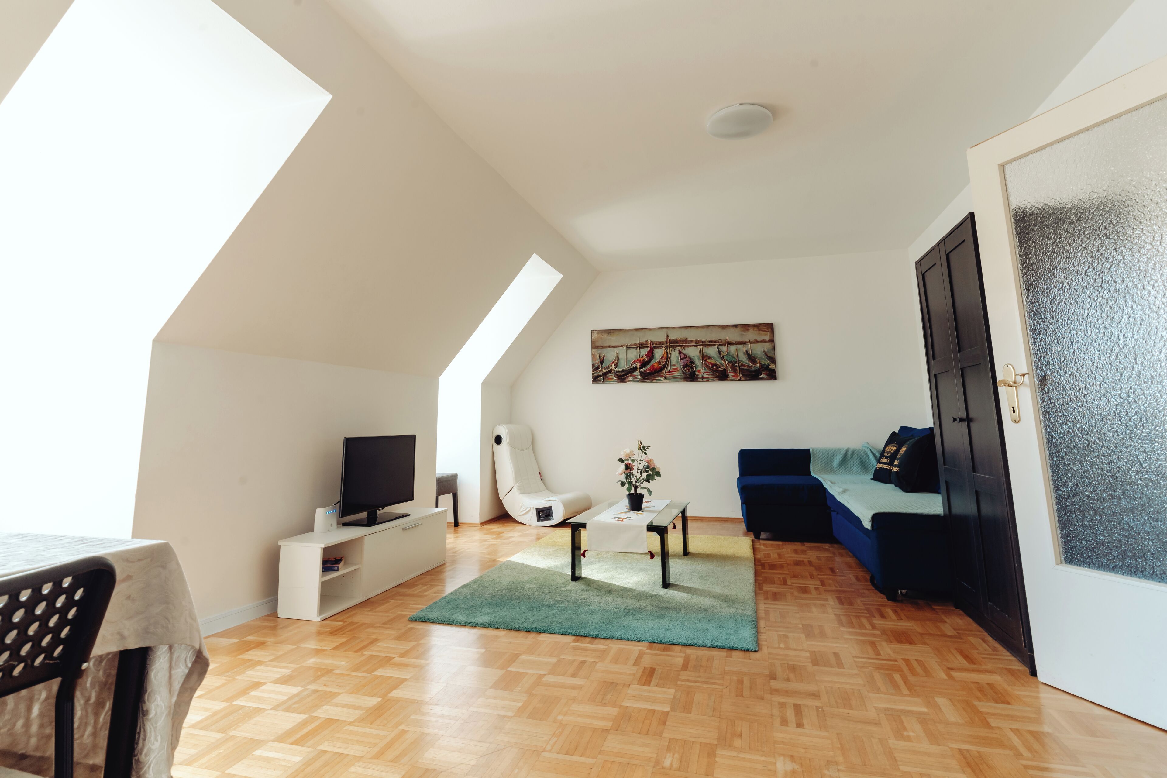 Een ruime woonkamer met een houten vloer, een witte bank, een blauwe bank, een klein tafeltje met een plant, een witte fauteuil en een televisie op een witte kast.