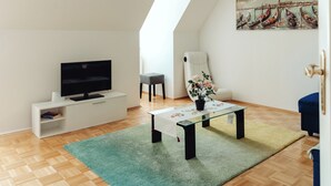A modern living room with a flat-screen TV, a glass coffee table, a potted plant, and a painting of boats on the wall. - Lilians Cosy Apartment Jakomini 3B (Graz)
