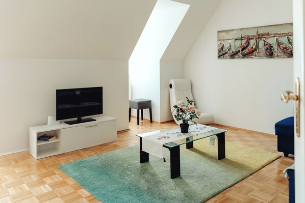 A modern living room with a flat-screen TV, a glass coffee table, a potted plant, and a painting of boats on the wall.