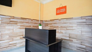 Un mostrador de recepción con un fondo de pared de piedra, un letrero que dice "KOOL KOST" y una planta en maceta en la pared.
