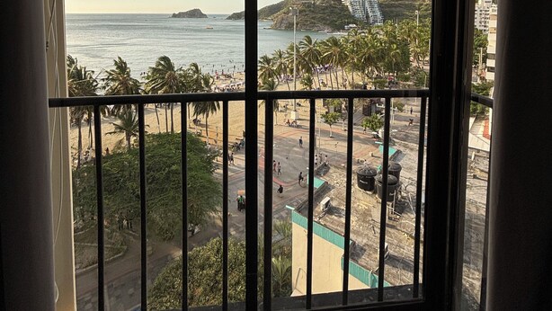 Vista desde una ventana con balcón sobre una playa, palmeras y una colina distante con un edificio.