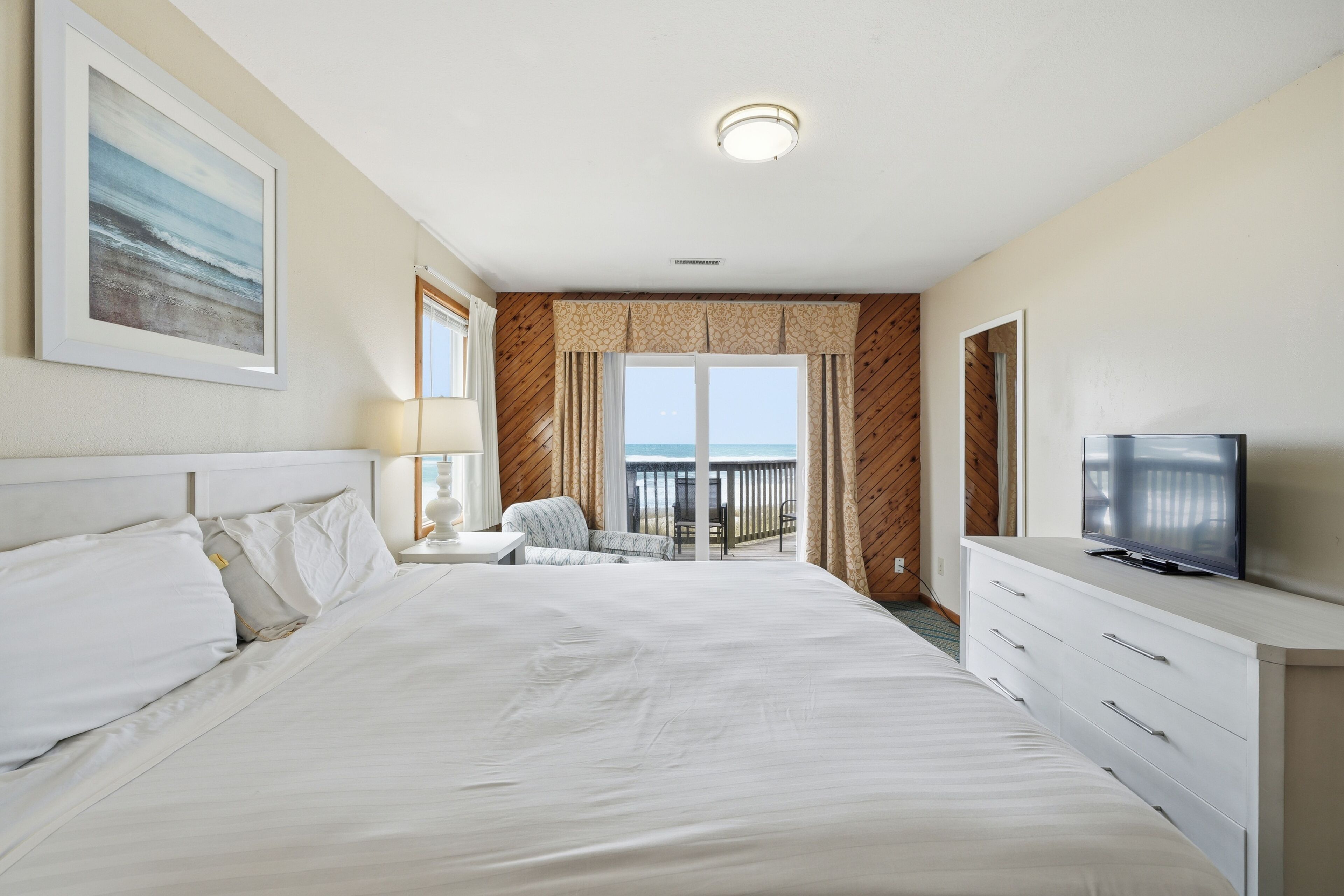 Ein Hotelzimmer mit einem großen Bett, einem Fernseher, einem Schreibtisch mit Lampe und Blick aufs Meer.