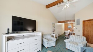 A living room with a flat-screen TV on a white cabinet, two patterned armchairs, a wooden ceiling fan, and a dining area with a table and chairs. - Charming Coastal-Facing 2-Bedroom Condo in Duck (Duck)