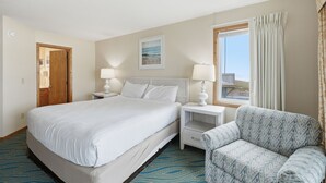A hotel room with a large bed, two bedside tables with lamps, a window with curtains, and a patterned armchair. - Charming Coastal-Facing 2-Bedroom Condo in Duck (Duck)