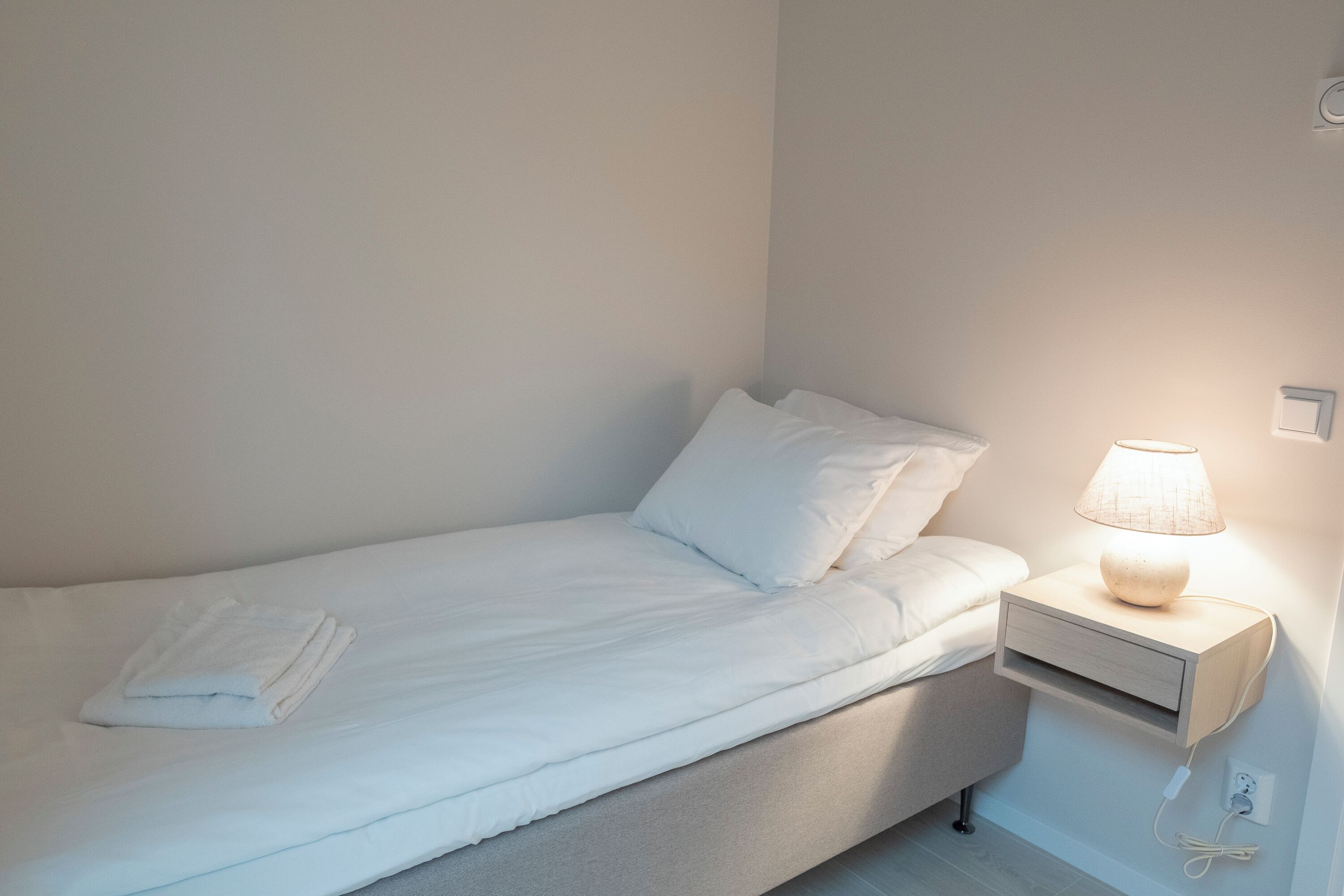 En enkeltseng med hvide sengetøj, et natbord med lampe, og en stikkontakt på væggen.