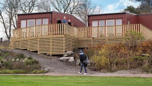 A wooden deck with a railing, a building with a red exterior, and a person walking on a path. - Skerike Golfhus (Vasteras)