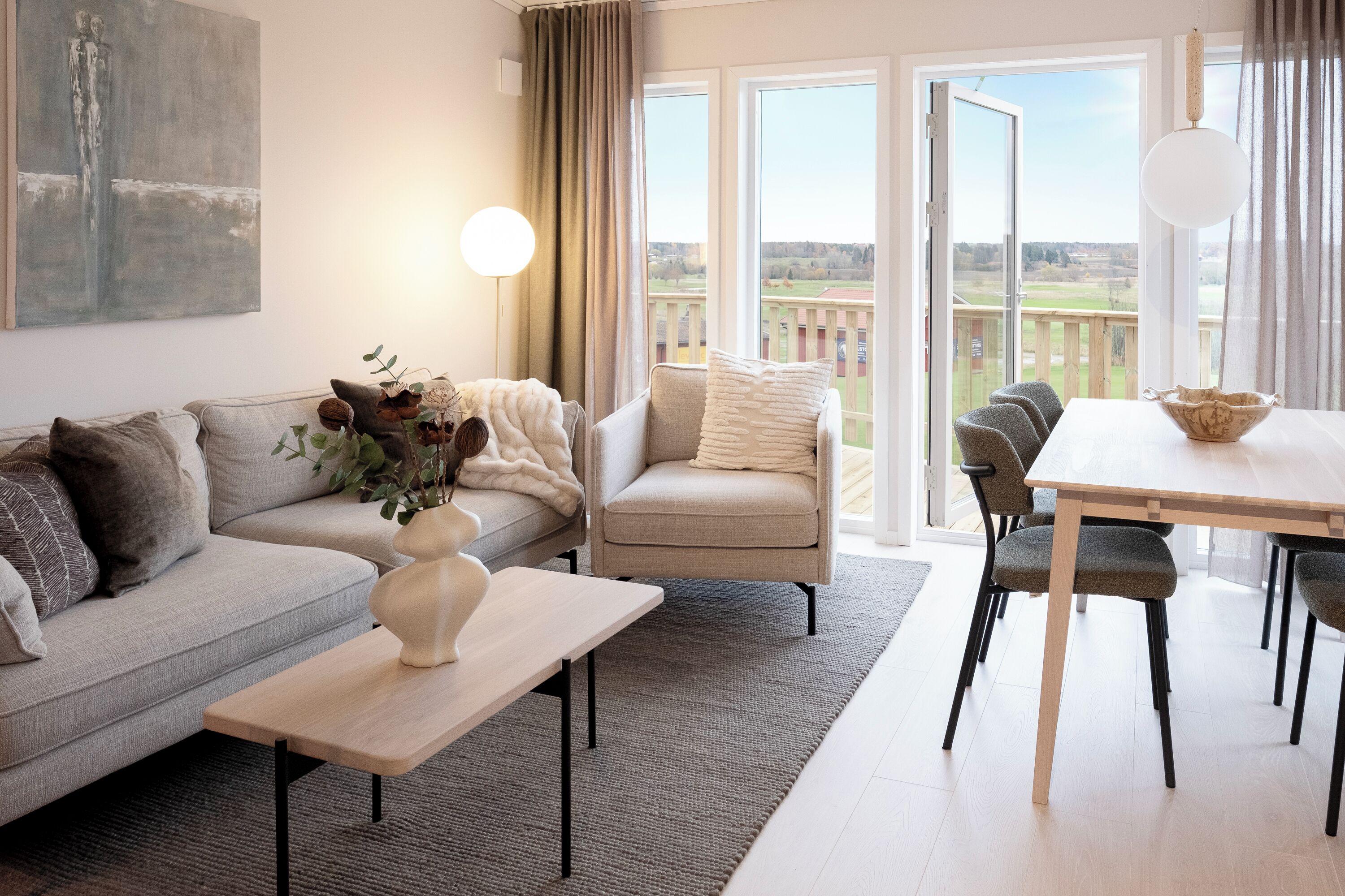 A modern living room with a sofa, armchair, dining table, and chairs. There is a painting on the wall and a view of the outdoors through large windows.