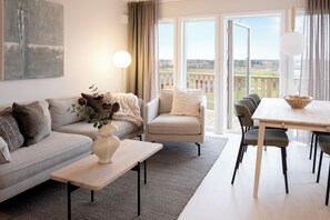 A modern living room with a sofa, armchair, dining table, and chairs. There is a painting on the wall and a view of the outdoors through large windows.