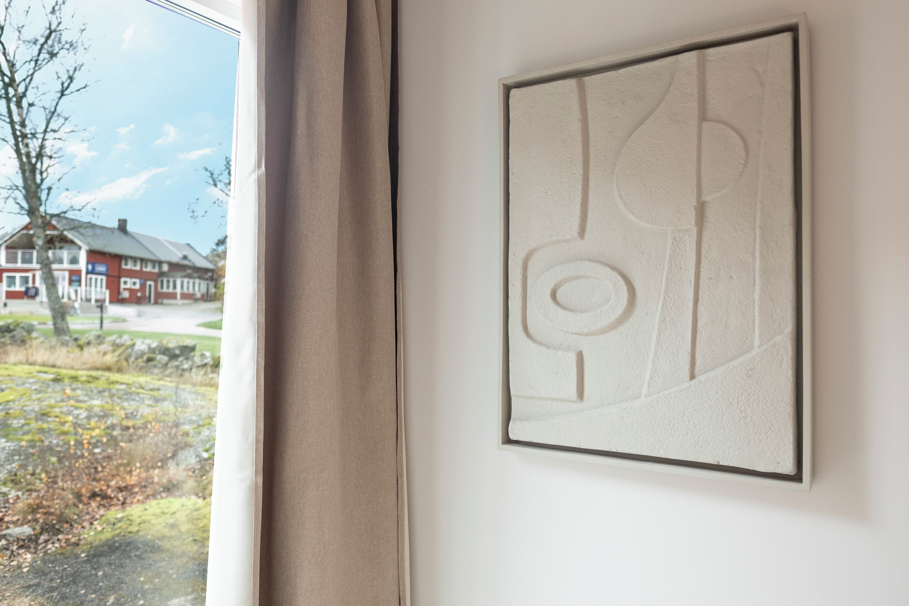A framed abstract artwork on a wall, a window with curtains, and a view of a house and trees outside.